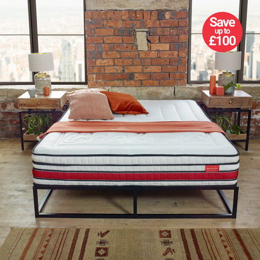 Mattress on a bed frame with decorative pillows in a room with brick walls and large windows.