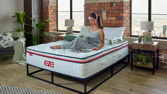 women sat on eze hybrid max mattress reading a magazine in a dressed bedroom with wooden floors and exposed brickwork with large windows looking out to a city skyline
