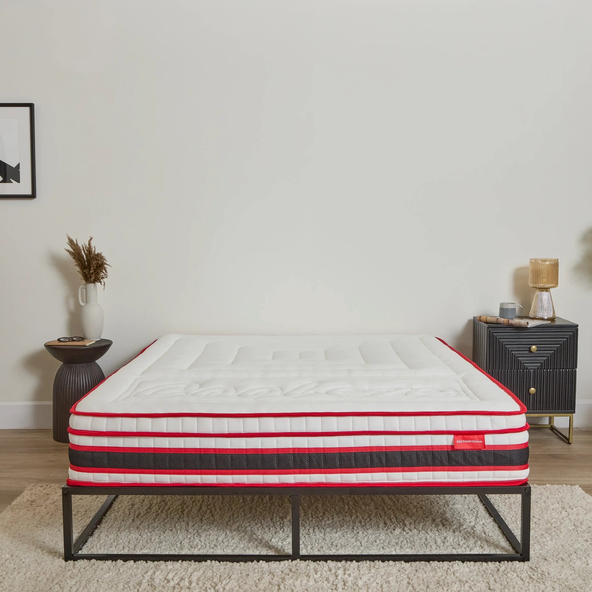 A mattress on a black metal bed frame in a styled bedroom setting with a white wall, side table, and lamp.