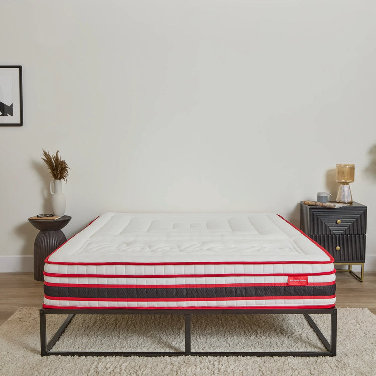 A mattress on a black metal bed frame in a styled bedroom setting with a white wall, side table, and lamp.