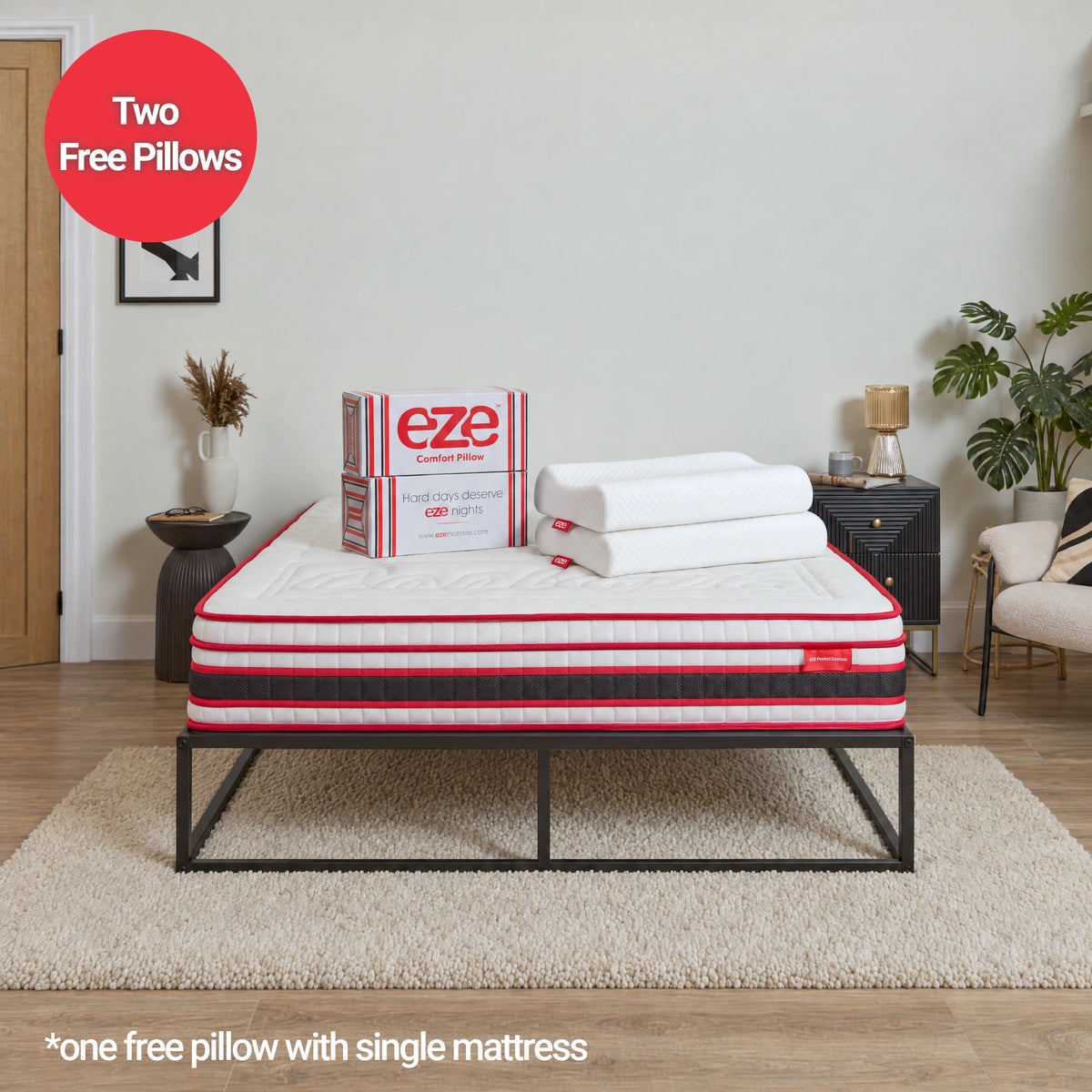 A mattress on a black metal bed frame in a styled bedroom setting, with a box and two folded white pillows on top. A red circular badge says 'Two Free Pillows' and text at the bottom says '*one free pillow with single mattress'.