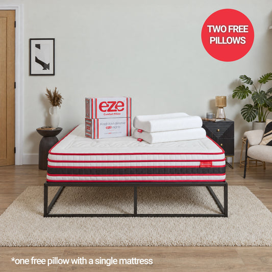 A mattress on a black metal bed frame in a styled bedroom setting, with a box and two folded white pillows on top. A red circular badge says 'TWO FREE PILLOWS' and text at the bottom reads '*one free pillow with a single mattress'.