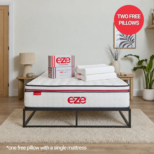 A white Eze mattress on a black metal bed frame in a styled bedroom setting, with two folded white pillows on top and an Eze product box beside it. A red circular badge says 'TWO FREE PILLOWS' and text at the bottom says '*one free pillow with a single mattress'.